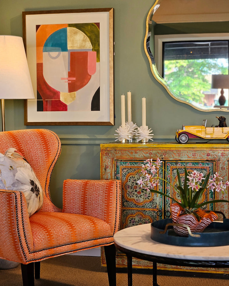A styled interior corner featuring a bright orange and pink striped wingback armchair, a colorful patterned console cabinet, a framed abstract art print on a sage green wall, and a round marble-top coffee table in the foreground.
