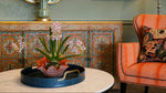 A styled interior corner featuring a vibrant orange armchair with a patterned accent pillow, a matching decorative cabinet, and a round marble coffee table holding a blue tray with a potted orchid.