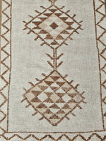 Close-up of a woven rug with a geometric diamond pattern in brown and beige on an off-white background.