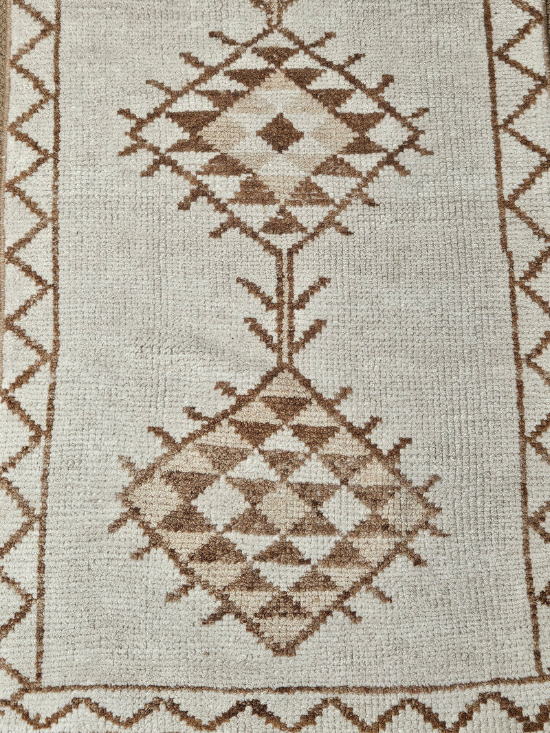 Close-up of a woven rug with a geometric diamond pattern in brown and beige on an off-white background.
