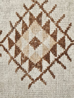 Close-up of a woven rug with a geometric diamond pattern in brown and beige on an off-white background.